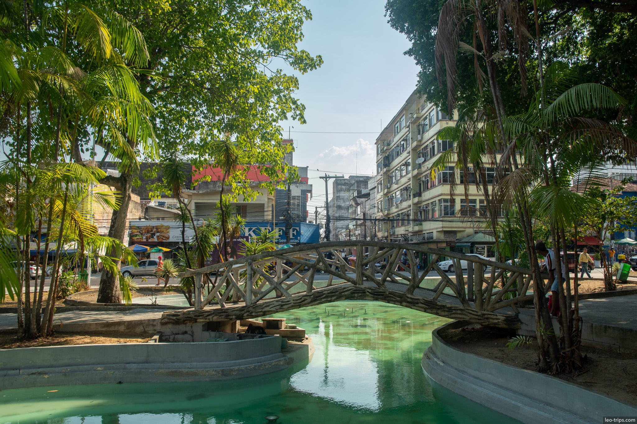 A rustic wooden footbridge crossing a green canal in a quiet, leafy urban park in Manaus