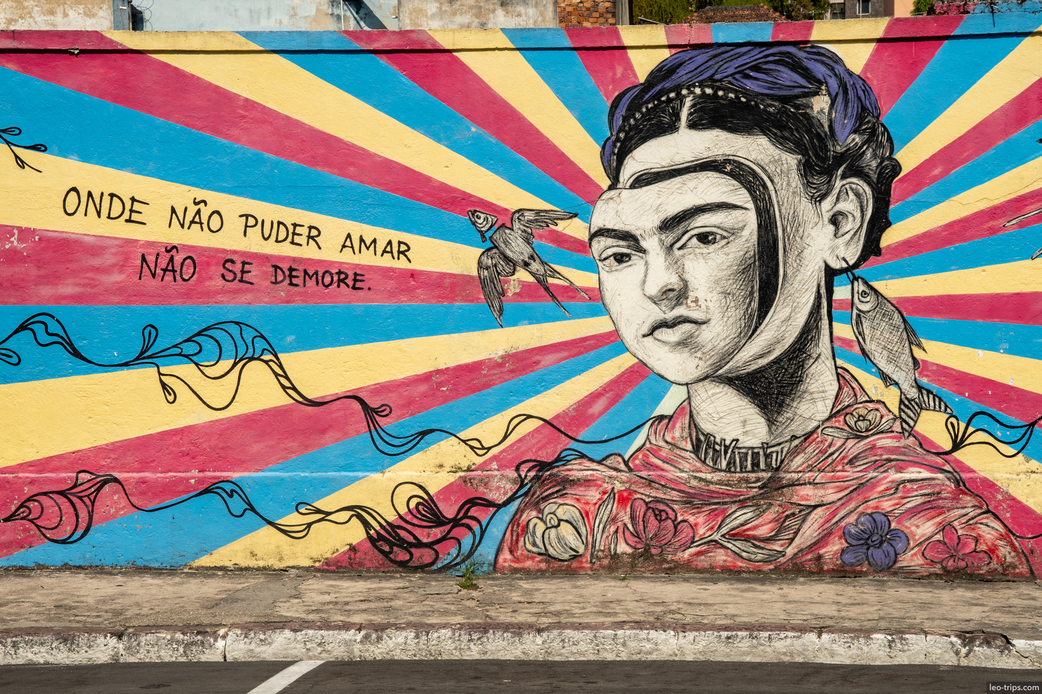 A vibrant street art mural depicting Frida Kahlo with the Portuguese quote 'Onde não puder amar, não se demore' (Where you cannot love, do not delay)