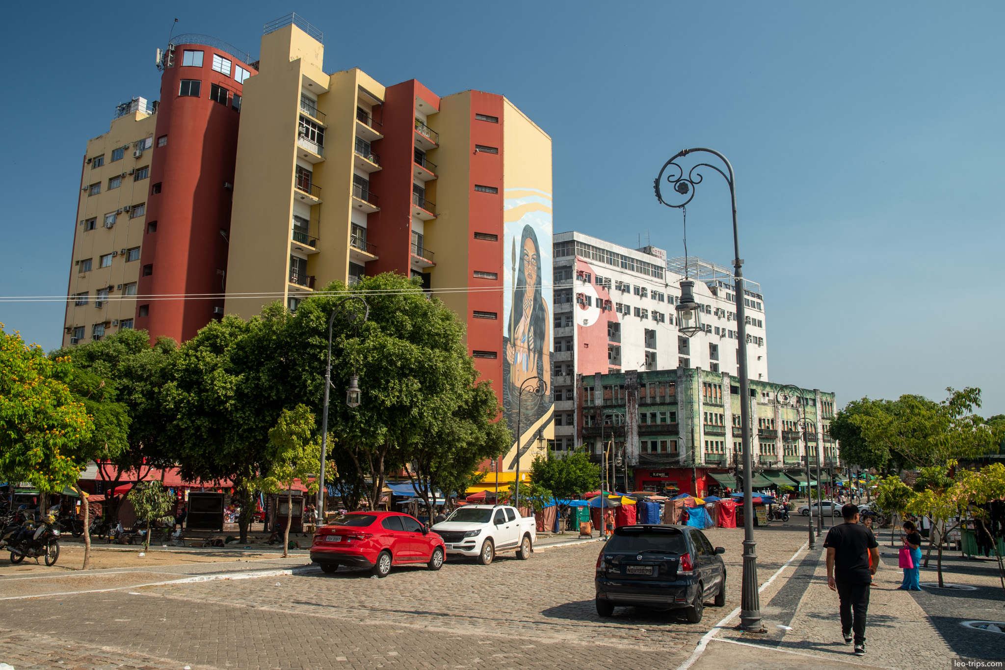 A sunny day at a public square in Manaus, showing a mix of trees, parking areas, and colorful modern apartment buildings