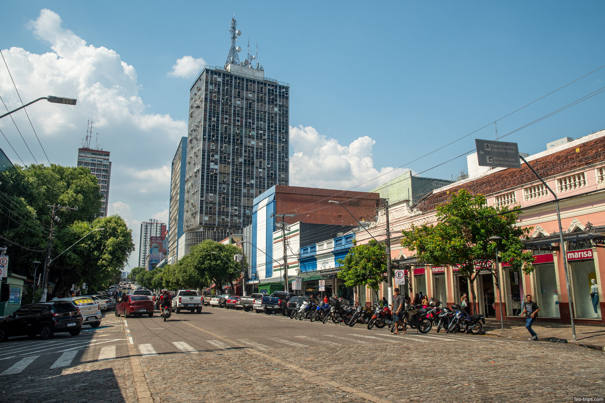 A perspective view of a busy downtown avenue in Manaus lined with trees, shops, and motorcycles