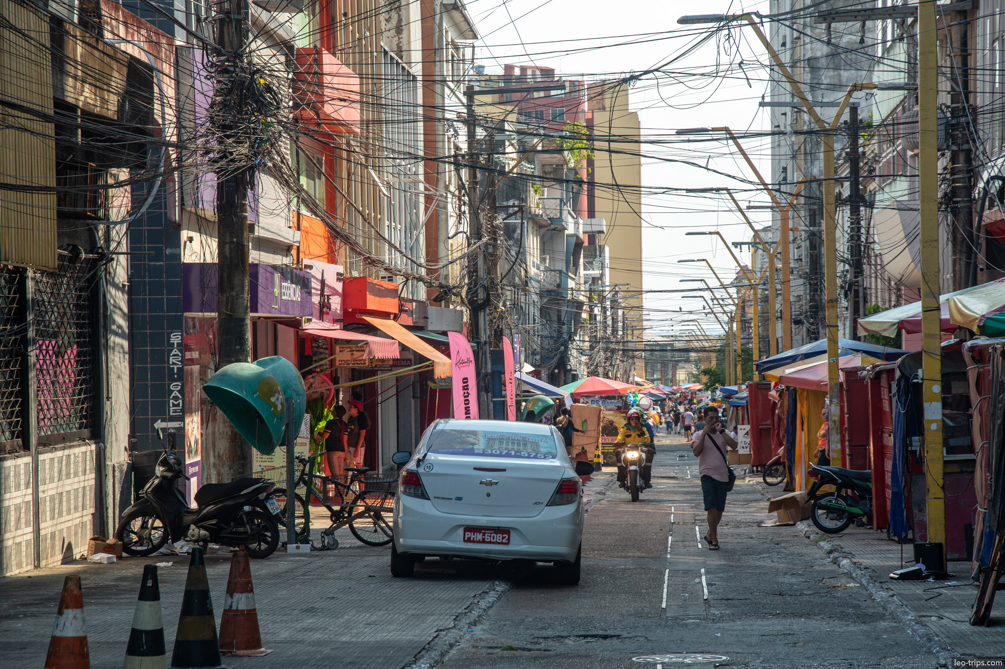 A narrow, busy street in the commercial district, cluttered with overhead wires and bustling with daily activity