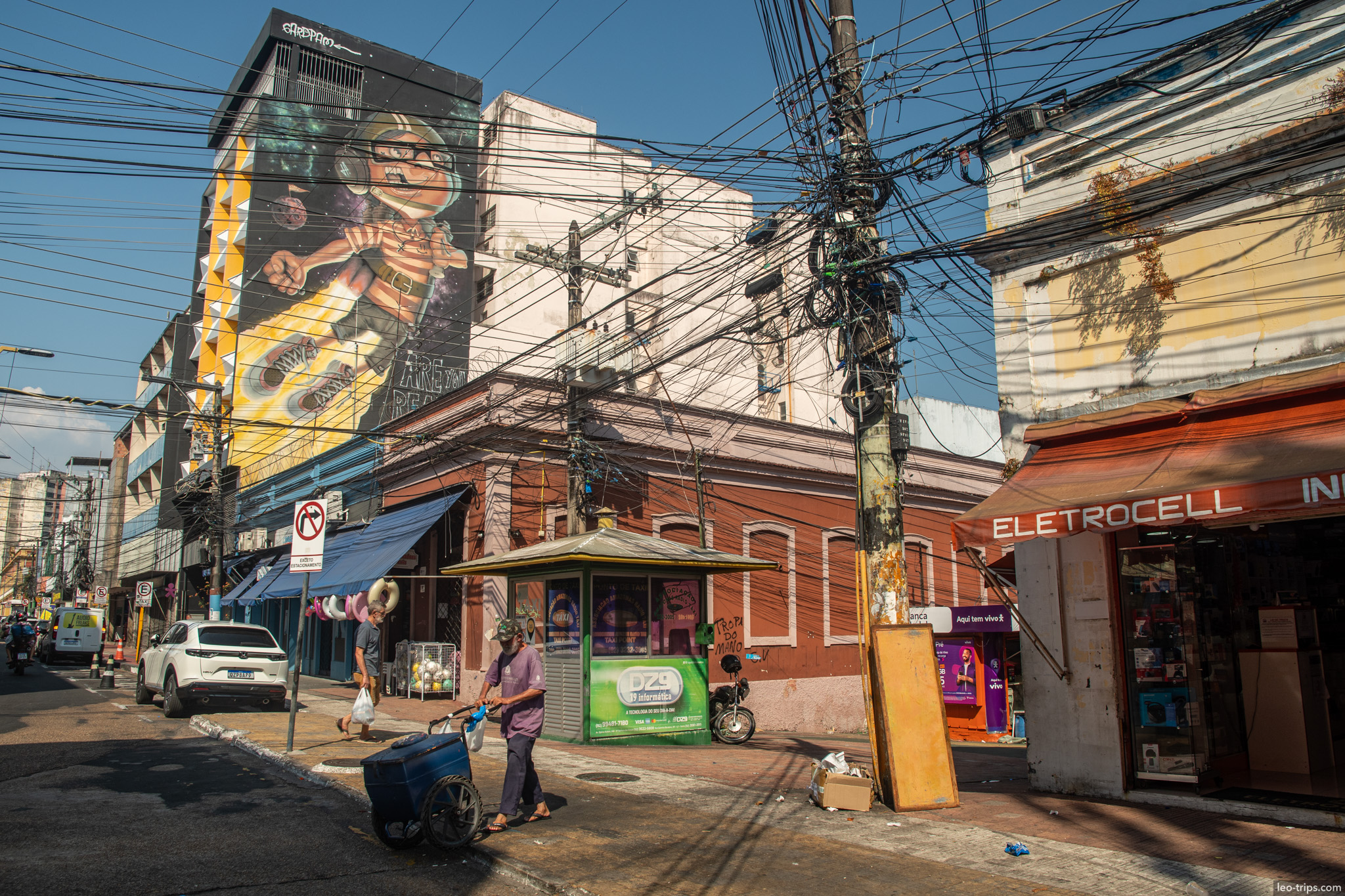 A street corner scene capturing the essence of the city: street vendors, tangled wires, and a large mural on the wall
